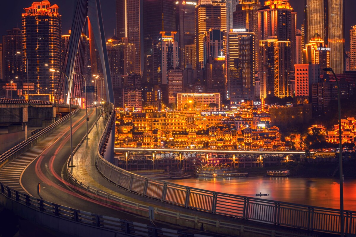 Noćni pogled na grad Chongqing s osvijetljenim neboderima u pozadini i modernim mostom koji prelazi rijeku, stvarajući dinamičan urbani pejzaž.