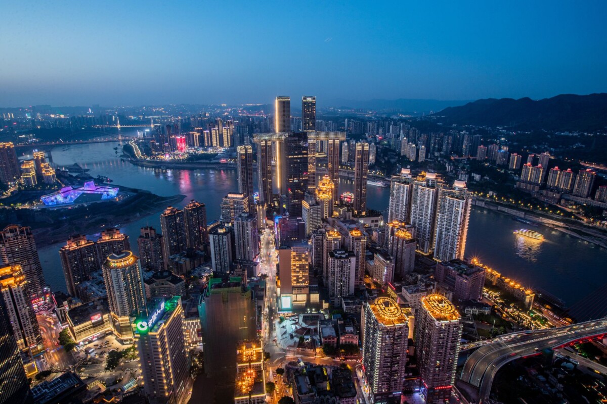 Panoramski pogled na grad Chongqing u sumrak, s visokim neboderima osvijetljenim gradskom rasvjetom, rijekama koje vijugaju kroz urbano područje i planinskim obrisima u pozadini.