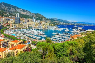 Panoramski pogled na Monte Carlo s luksuznom marinom punom jahti, modernim neboderima, zelenilom u prvom planu i planinama u pozadini.