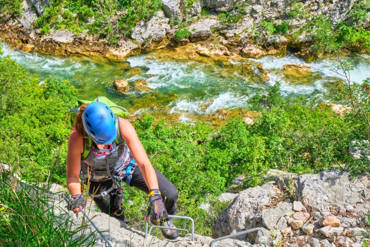 kanjon-rijeke-čikole-zagora Penjačica s kacigom i zaštitnom opremom uspinje se stjenovitim putem iznad rijeke Čikole, koja vijuga kroz zeleni kanjon s kristalno čistom vodom.