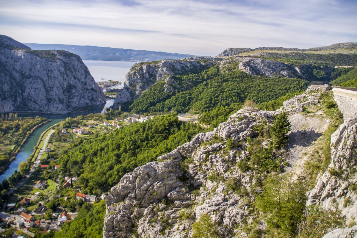 Pogled na Omiš, grad smješten između stijena i ušća rijeke Cetine, s brdima, morem u daljini i zelenilom koje okružuje grad.