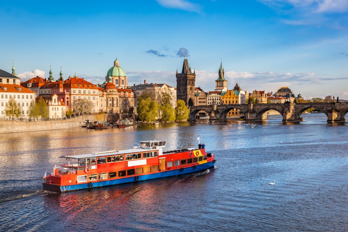 Rijeka Vltava u Pragu s crveno-bijelim turističkim brodom u prvom planu. U pozadini se proteže povijesni Karlov most s gotičkim tornjevima, okružen šarenim baroknim i renesansnim građevinama starog grada pod vedrim nebom.