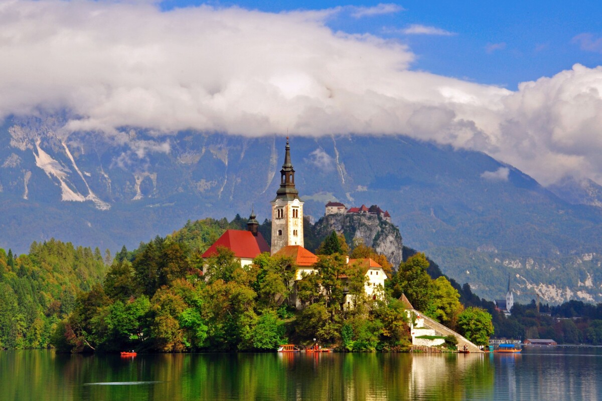Otočić na Bledskom jezeru s crkvom Uznesenja Marijina, okružen jesenskim bojama i planinama u pozadini – čarobna slovenska destinacija u srcu prirode. Savršen izlet na Bled.