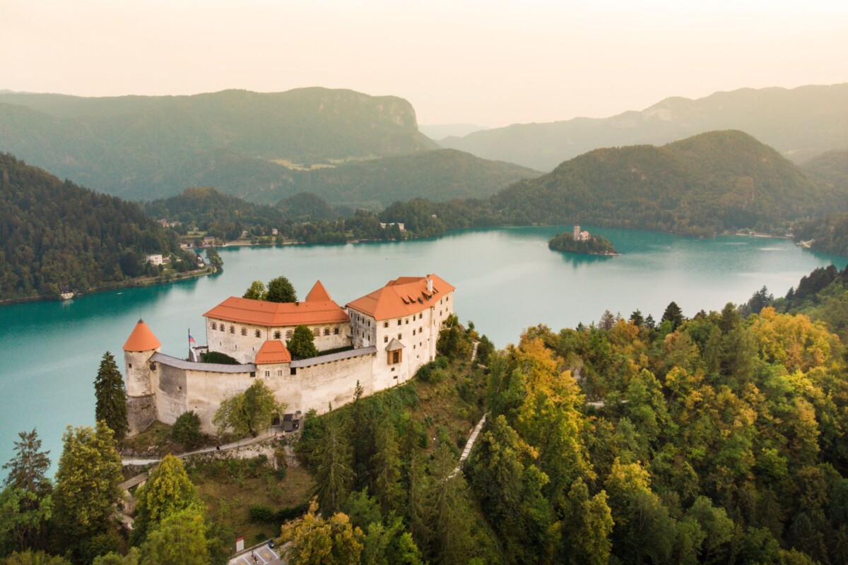 bledski-dvorac-s-pogledom-na-jezero Bledski dvorac iz zraka s pogledom na Bledsko jezero i otočić – povijesna znamenitost Slovenije smještena na litici iznad jezera, okružena alpskim krajolikom.