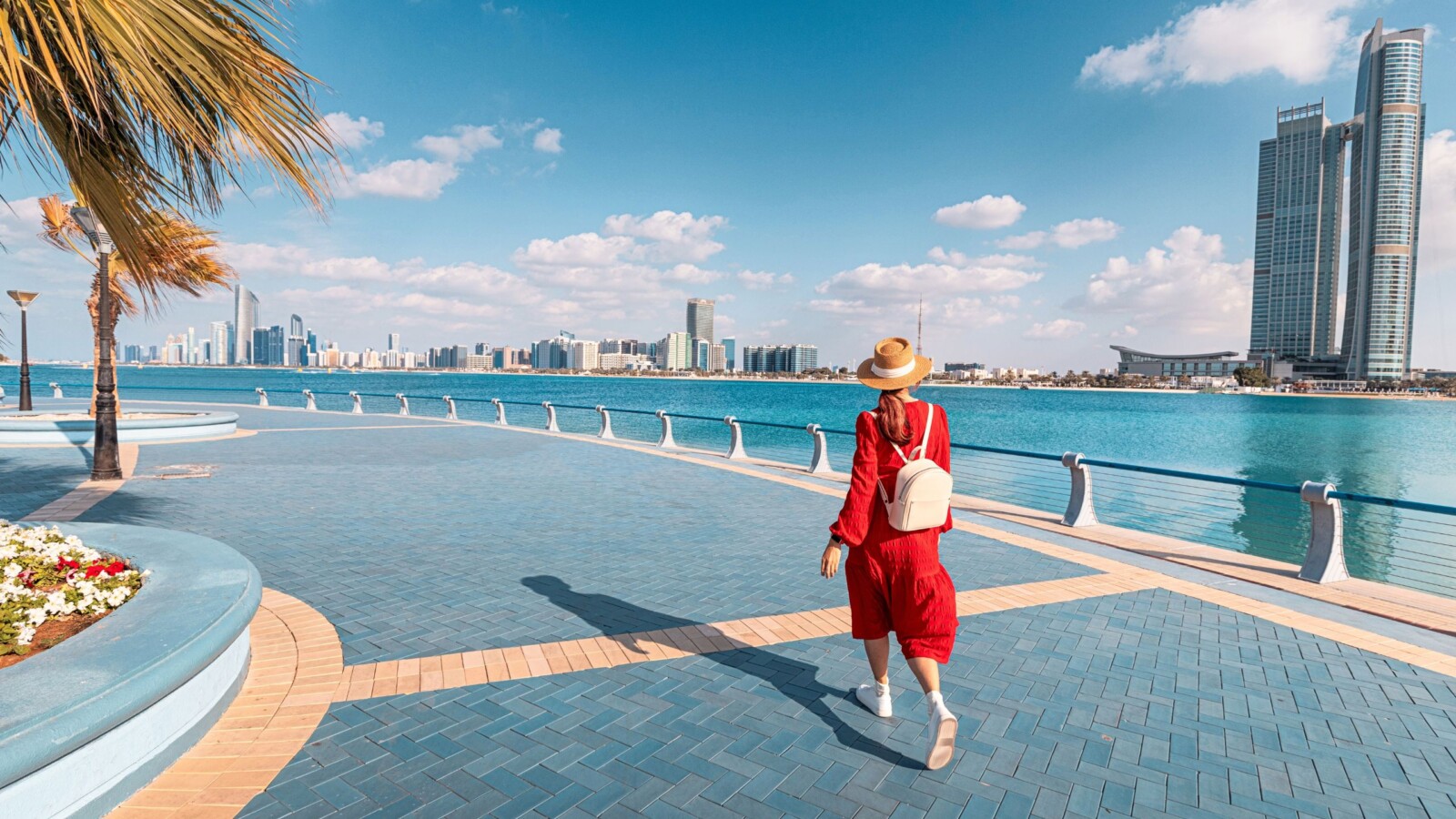 abu-dhabi-setnica-uz-more Žena u crvenoj haljini šeta Corniche šetnicom u Abu Dhabiju uz more i panoramu grada pod vedrim nebom