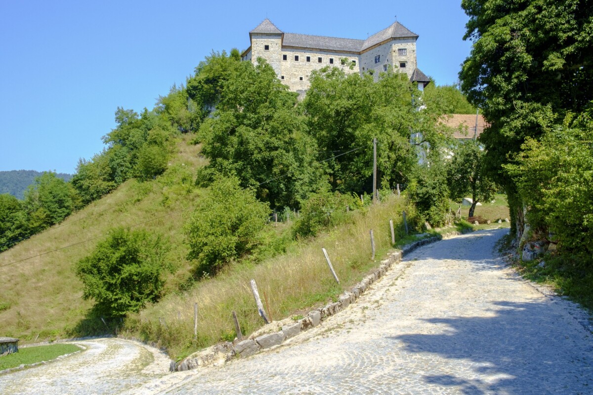 Pogled na dvorac Kostel u Sloveniji, smješten na uzvisini iznad popločane seoske ceste, okružen zelenilom i šumom pod vedrim plavim nebom.