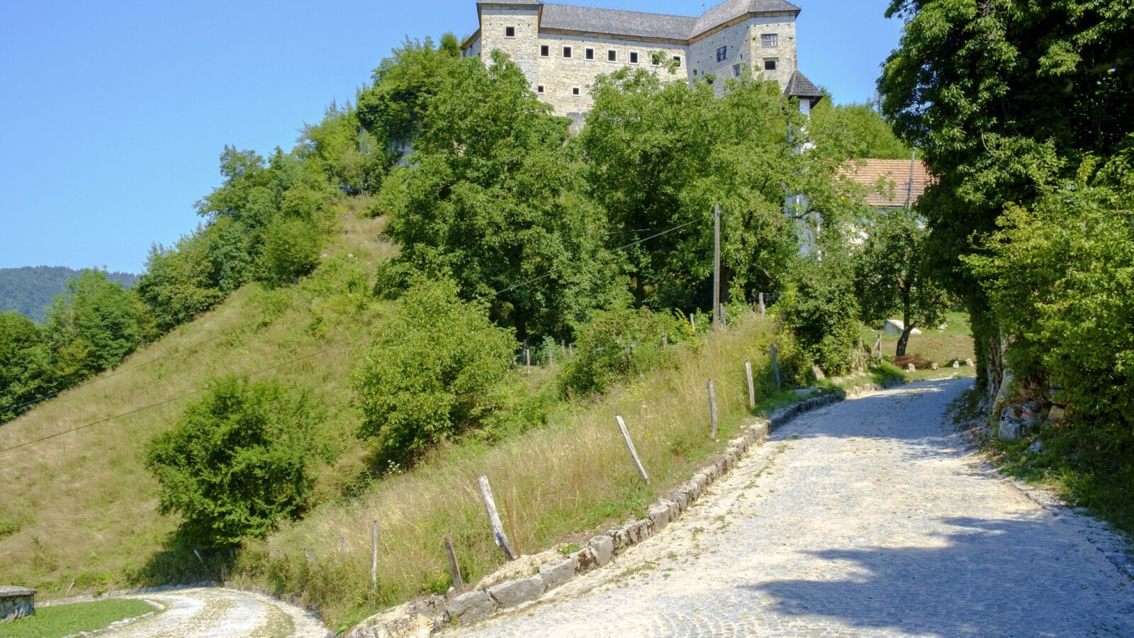 Pogled na dvorac Kostel u Sloveniji, smješten na uzvisini iznad popločane seoske ceste, okružen zelenilom i šumom pod vedrim plavim nebom.