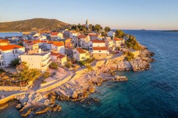 Pogled iz zraka na Primošten, slikoviti dalmatinski gradić smješten na poluotoku, s kamenim kućama crvenih krovova, crkvom na uzvisini i okruženjem tirkiznog Jadranskog mora u zlatnoj svjetlosti zalaska sunca.