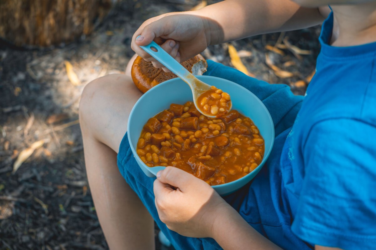 Osoba sjedi na otvorenom i drži zdjelu s grahom i kobasicom te jede drvenom žlicom – tradicionalni obrok za Praznik rada.