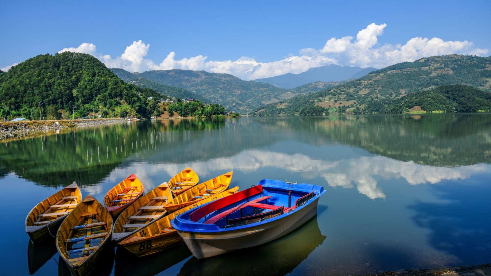 Čamci na mirnoj površini jezera Begnas u Pokhari, Nepal, okruženi planinama i plavim nebom