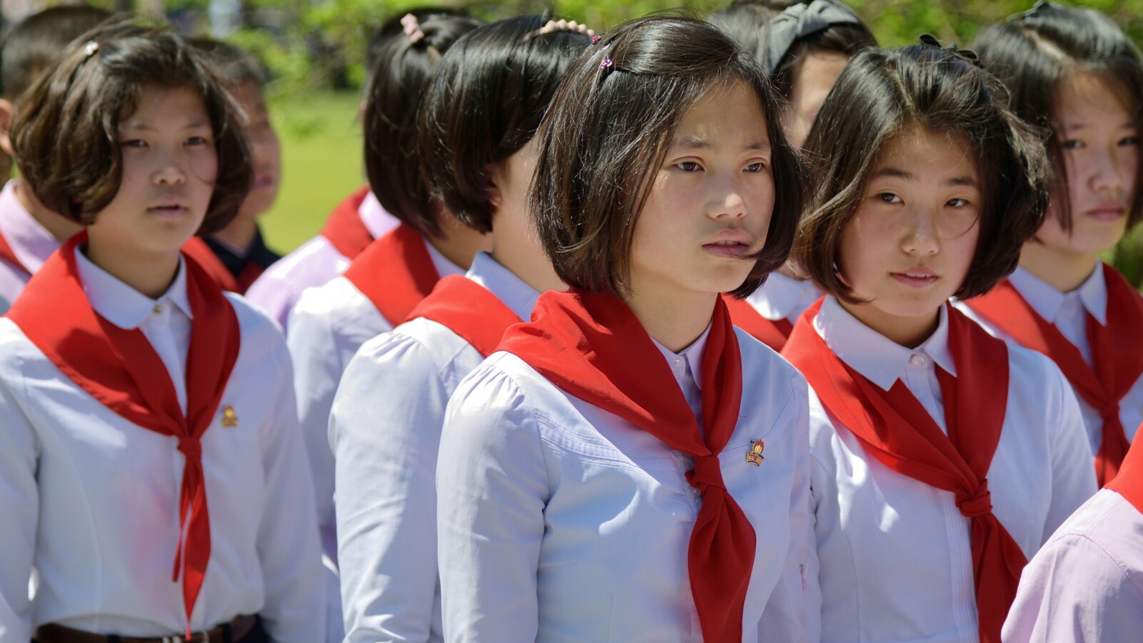 ljudi-u-sjevernoj-koreji Skupina sjevernokorejskih učenica u školskim uniformama s crvenim maramama i uredno podšišanim frizurama, stoje na otvorenom tijekom službenog događaja