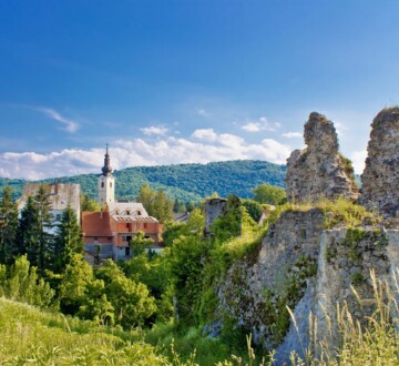 Pogled na staru utvrdu i crkvu u Slunju okružene zelenilom, uz planine u pozadini pod vedrim nebom. AKo poželite otići na izlet u Slunj, ovaj grad neće vas razočarati.