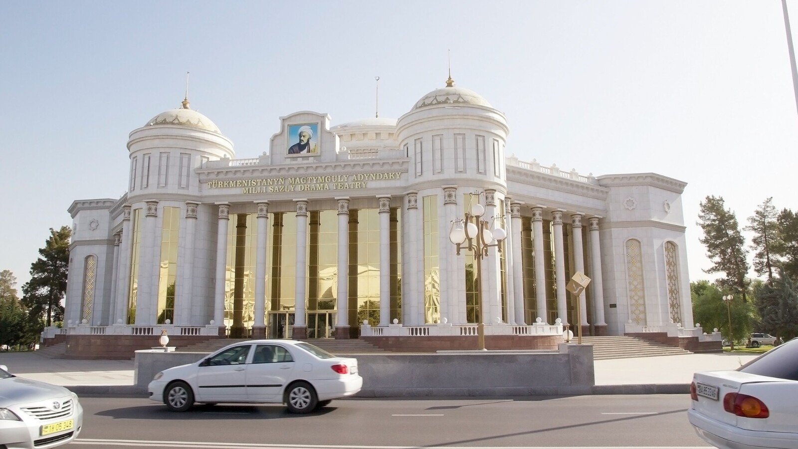 Bijela zgrada s kupolama i zlatnim staklima u Ašgabatu, ispred koje vozi bijeli automobil, simbol stroge estetike glavnog grada Turkmenistana.