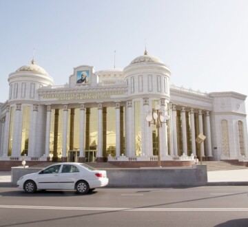 Bijela zgrada s kupolama i zlatnim staklima u Ašgabatu, ispred koje vozi bijeli automobil, simbol stroge estetike glavnog grada Turkmenistana.