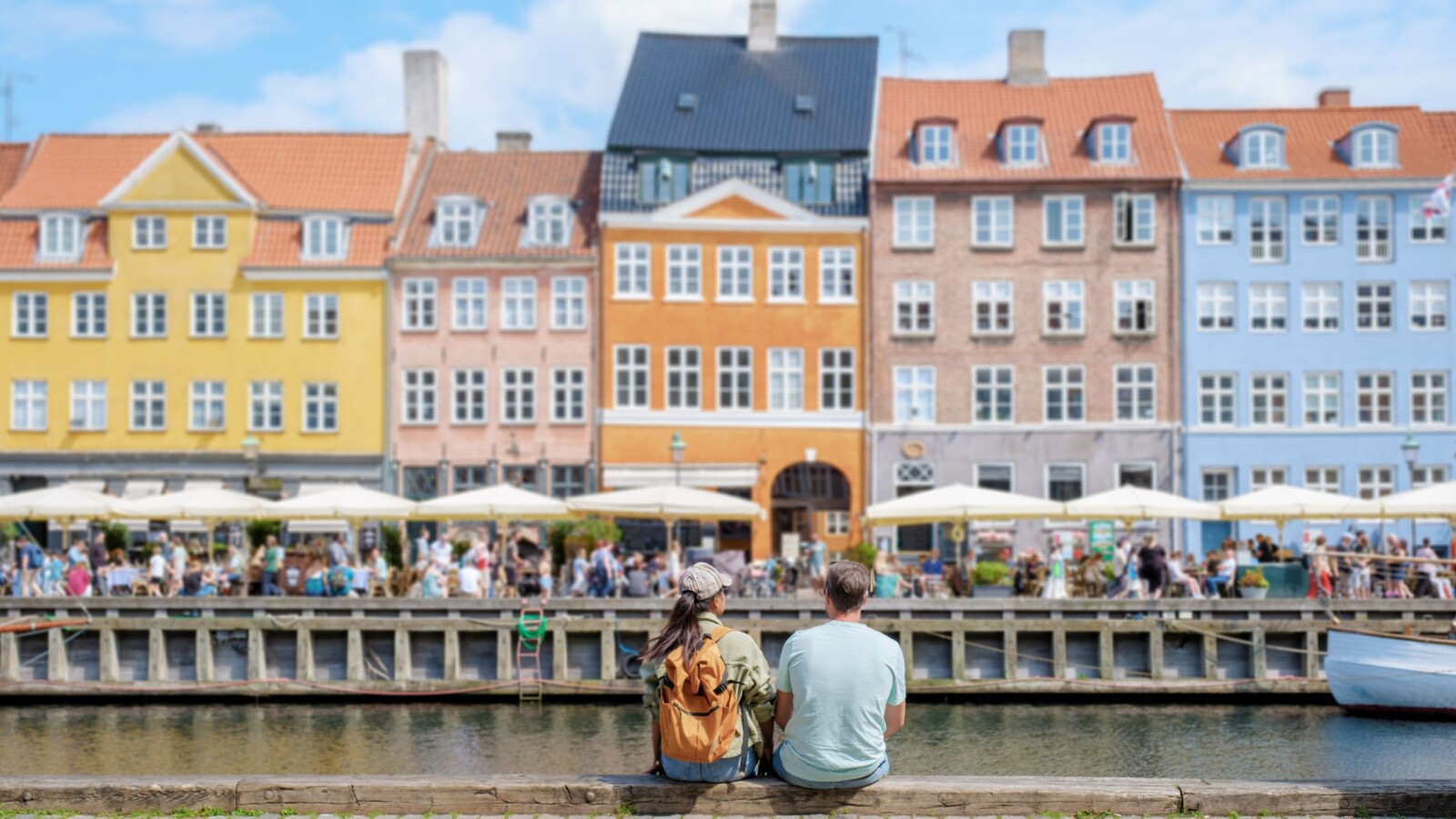 kopenhagen-glavni-grad-danske Dvoje sretnih ljudi sjedi uz kanal u Kopenhagenu s pogledom na šarene povijesne zgrade i živahne terase kafića u pozadini. Kopengahen koji slovi za najsretniji grad na svijetu.