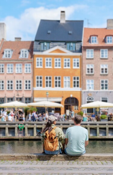 kopenhagen-glavni-grad-danske Dvoje sretnih ljudi sjedi uz kanal u Kopenhagenu s pogledom na šarene povijesne zgrade i živahne terase kafića u pozadini. Kopengahen koji slovi za najsretniji grad na svijetu.