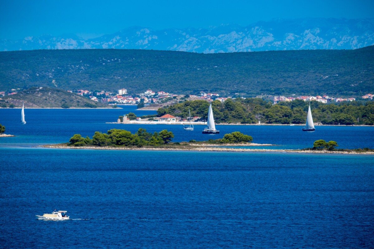 Pogled na otok Babac u Pašmanskom kanalu s jedrilicama i obalom u pozadini.