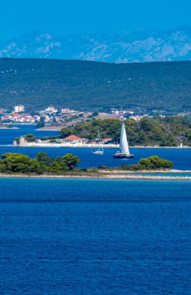 Pogled na otok Babac u Pašmanskom kanalu s jedrilicama i obalom u pozadini.