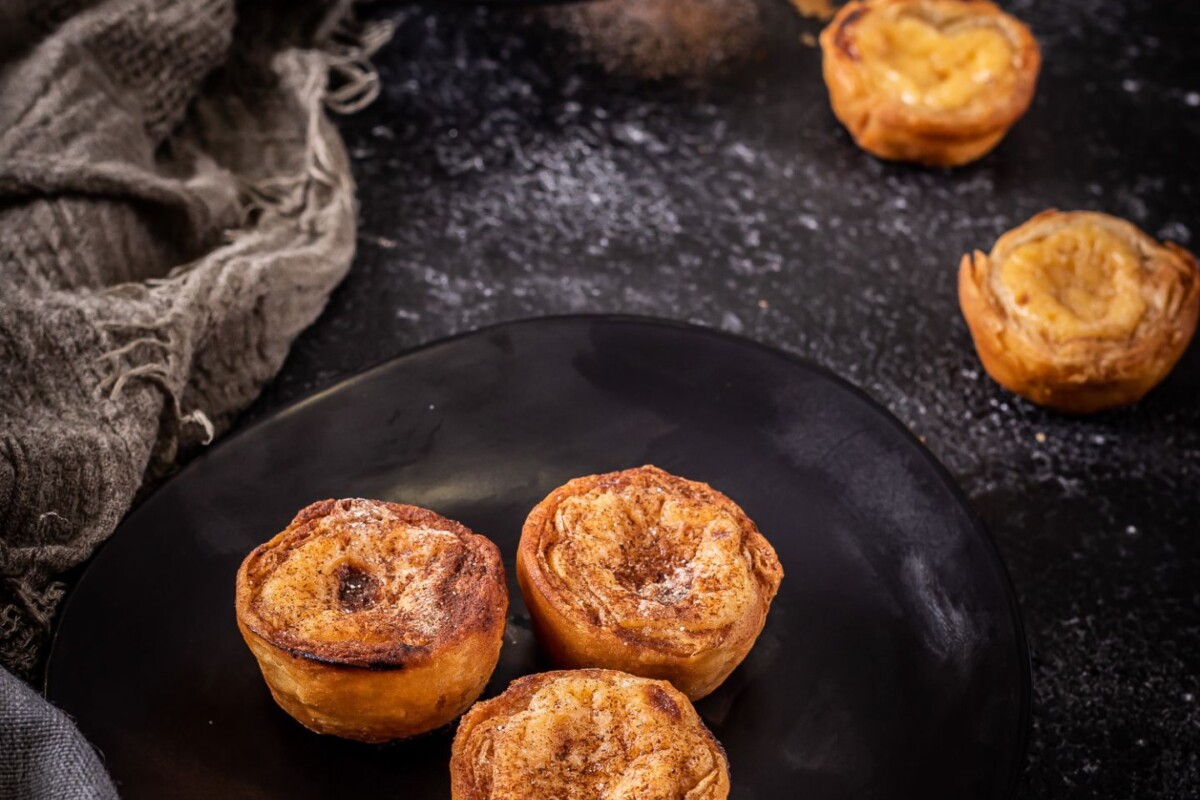 pastel-de-nata-1 Pastel de Nata, poznat i kao Pasteis de nata – tradicionalna portugalska slastica s hrskavim lisnatim tijestom i kremastim punjenjem, poslužena na crnom tanjuru uz rustikalnu pozadinu. Savršen recept za Pastel de Nata kod kuće.