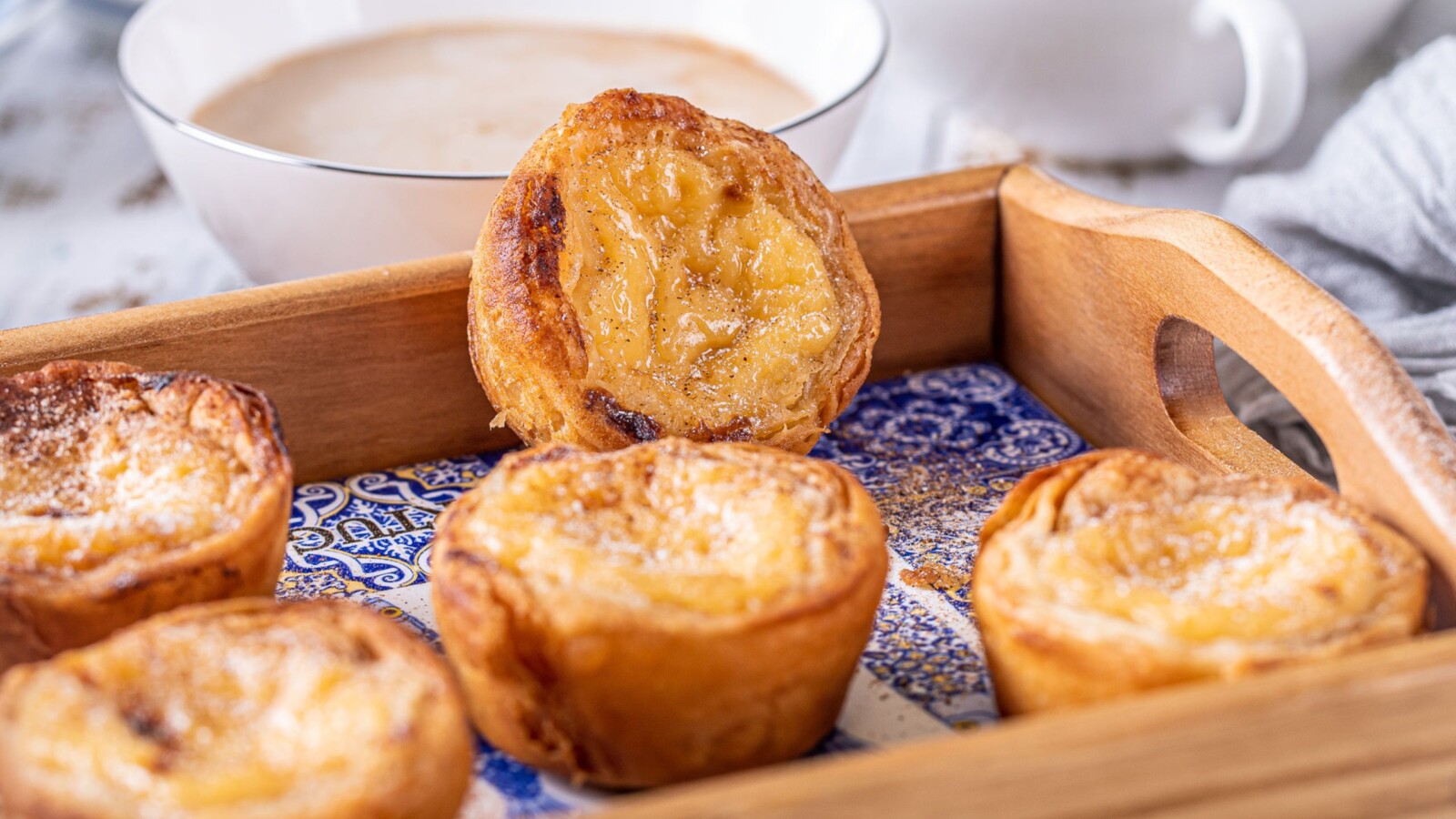 Pastel de Nata – tradicionalni portugalski kolačići od lisnatog tijesta s kremastim punjenjem, posluženi na drvenom pladnju s uzorkom pločica i kavom u pozadini.