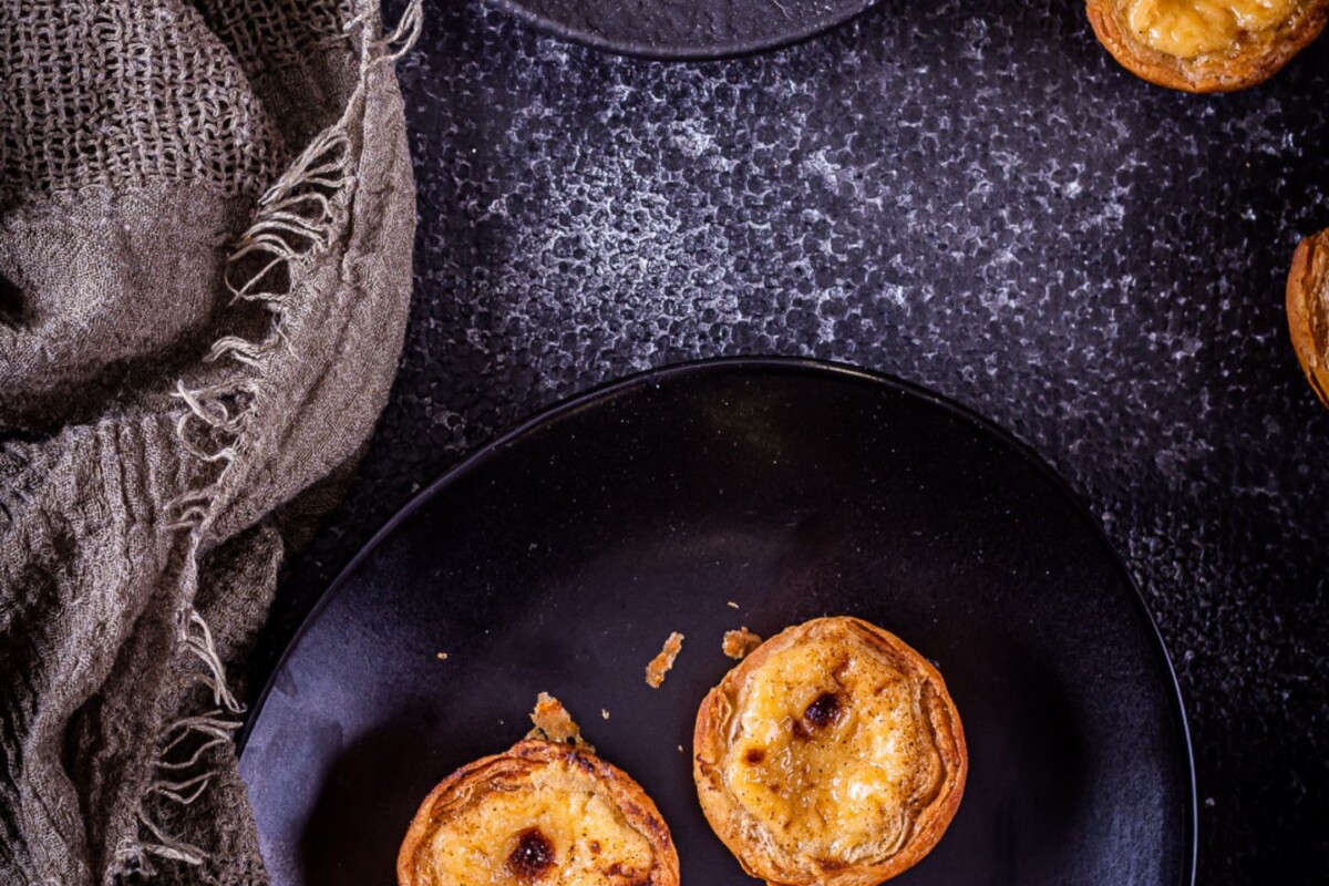pastel-de-nata-priprema-8 Tri Pastel de Nata (Pasteis de nata) kolačića na crnom tanjuru s tamnom rustikalnom pozadinom i cimetom u pozadini – tradicionalni portugalski Pasteis de nata po domaćem receptu.