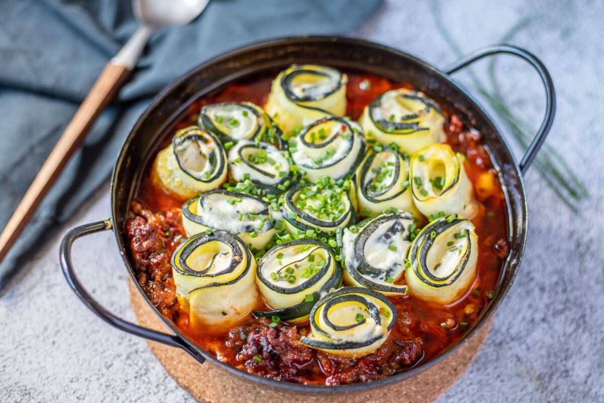 Punjene rolice od tikvica u umaku od mljevenog mesa poslužene u crnoj posudi, zapečene do zlatne boje i posute svježim vlascem.