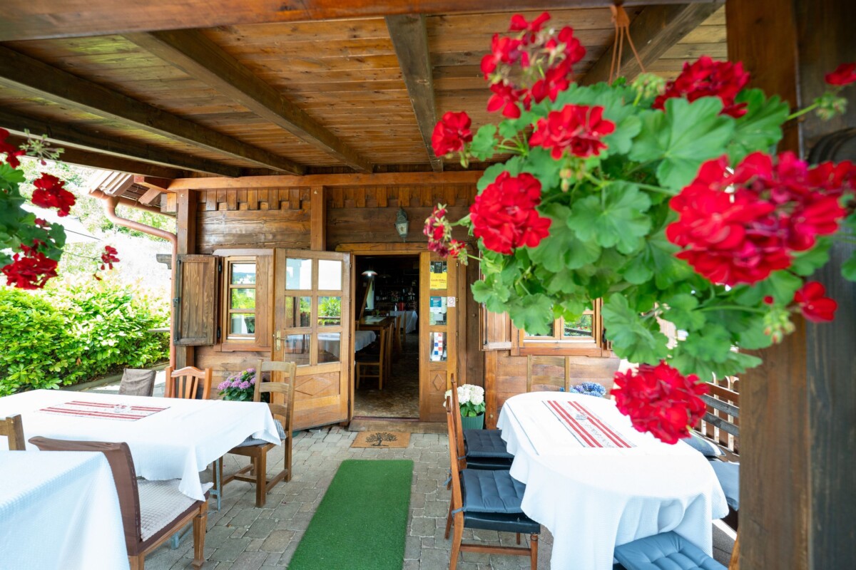 Ulaz u restoran Karlo na Plešivici s drvenom terasom, stolovima prekrivenim bijelim stolnjacima i visećim crvenim pelargonijama u prvom planu.