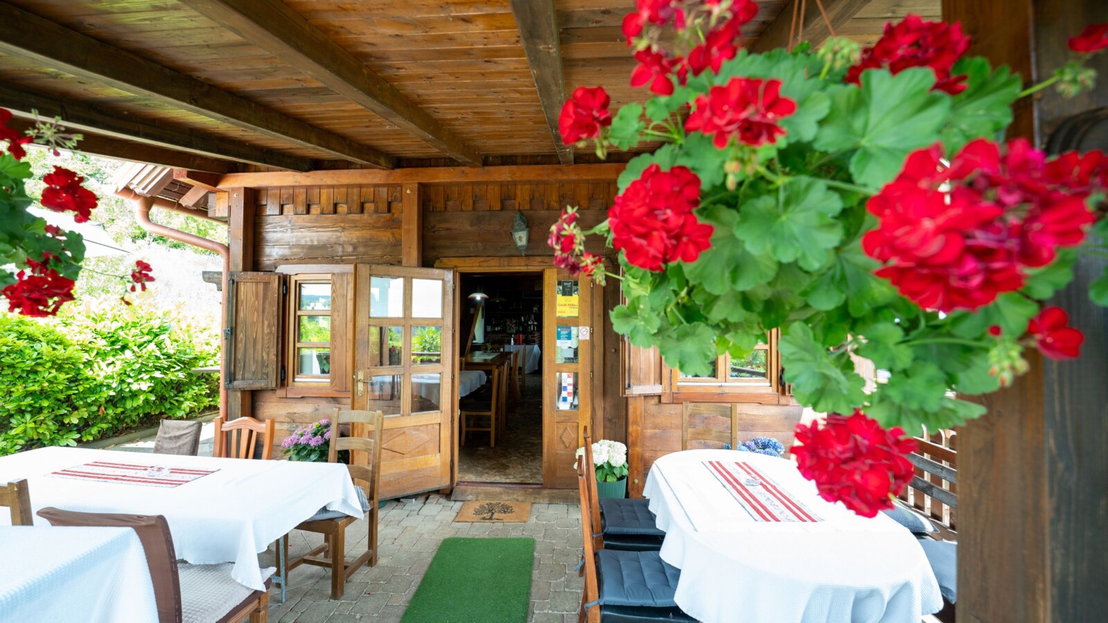 restoran-karlo-plesivica Ulaz u restoran Karlo na Plešivici s drvenom terasom, stolovima prekrivenim bijelim stolnjacima i visećim crvenim pelargonijama u prvom planu.