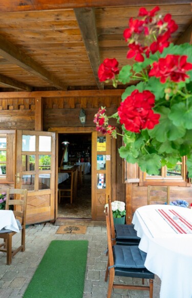 Ulaz u restoran Karlo na Plešivici s drvenom terasom, stolovima prekrivenim bijelim stolnjacima i visećim crvenim pelargonijama u prvom planu.