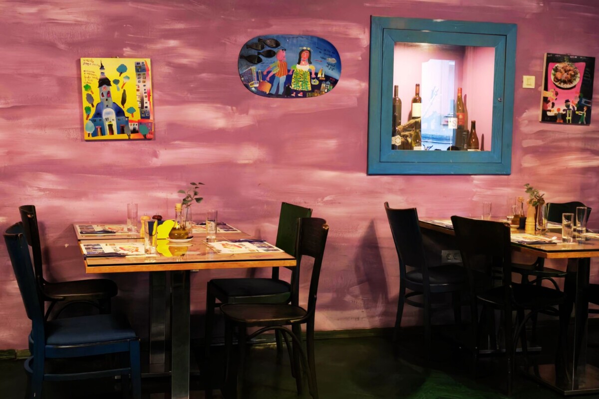 ribica-i-tri-tockice-interijer Detalj interijera restorana Ribica i tri točkice s ljubičastim zidovima, umjetničkim ilustracijama i drvenim stolovima pripremljenima za goste.