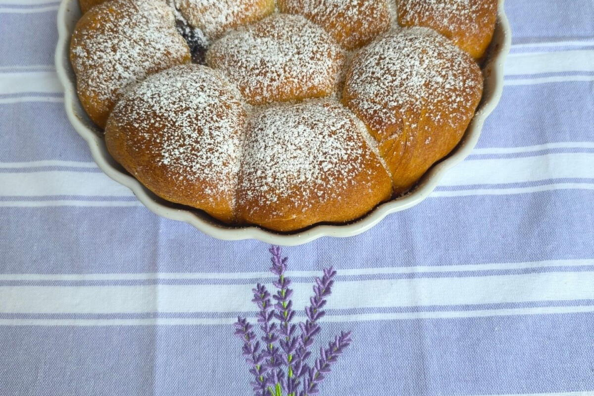 Pečene buhtle posute šećerom u prahu, složene u okruglu posudu, s jednom prerezanom buhtlom koja otkriva punjenje od džema. Postavljeno na stolnjaku s motivom lavande.