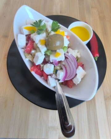 Gotova šopska salata u bijeloj zdjeli s feta sirom, povrćem i maslinom, poslužena s maslinovim uljem na crnom tanjuru.“