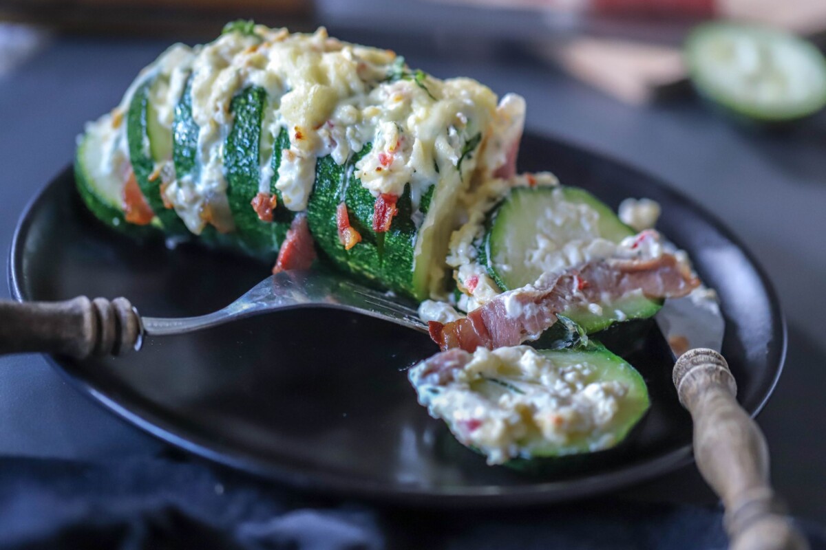 Sočne Hasselback tikvice punjene sirom, slaninom i začinima, poslužene na crnom tanjuru s rustikalnim priborom za jelo. Recept za hasselback tikvice.