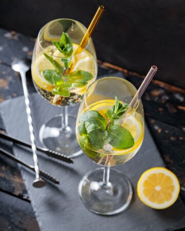 Dvije čaše koktela Hugo Spritz sa slamkama, svježom mentom, limunom i ledom, poslužene na tamnoj podlozi uz metalne barske alate i pola limuna za ukras.