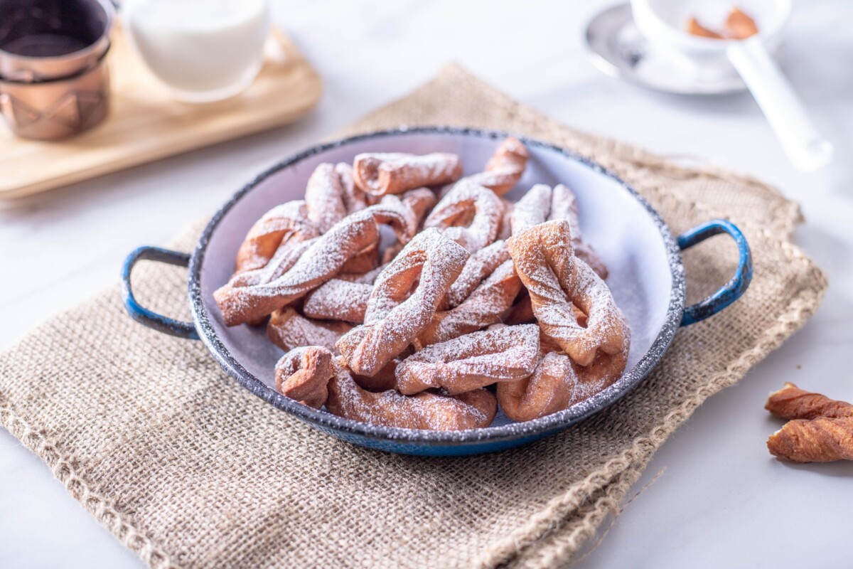 Kroštule posute šećerom u prahu servirane u bijeloj zdjeli s plavim rubom, na rustikalnoj podlozi od jute.