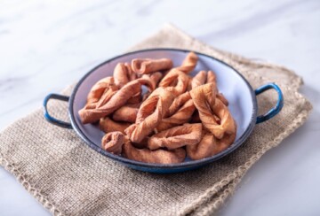 Svježe pržene kroštule bez šećera u prahu, poslužene u bijeloj zdjeli s plavim rubom na podlozi od jute, snimljene na svijetloj podlozi. Recept za kroštule.