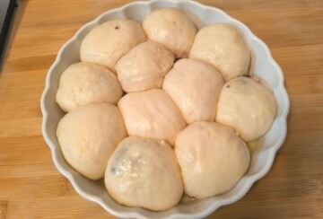 Sirove buhtle posložene u okruglu keramičku posudu, spremne za pečenje, na drvenoj radnoj površini. Recept za najmekše i najbolje buhtle.