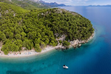 Zračni pogled na plažu Velika Duba u Živogošću, okruženu gustim borovima i kristalno čistim tirkiznim morem, s malim brodom usidrenim uz obalu.