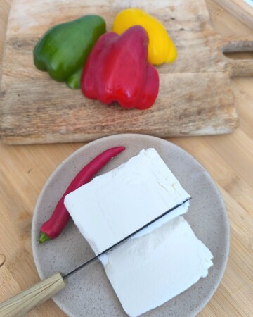 Priprema šopske salate: feta sir s papričicom na tanjuru uz nož, u pozadini svježe paprike u tri boje na drvenoj dasci.
