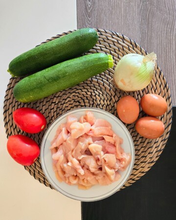 Sirovi sastojci za složenac od tikvica i piletine prikazani na podlošku: tikvice, luk, rajčice, jaja i nasjeckana pileća prsa u zdjeli.