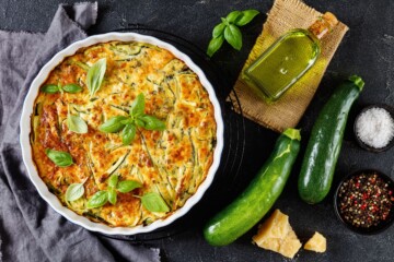 scarpaccia-italija-toskana Pečena scarpaccia s tikvicama i svježim bosiljkom u bijelom keramičkom kalupu, okružena tikvicama, maslinovim uljem i komadićima parmezana. Talijanska pita od tikvica.