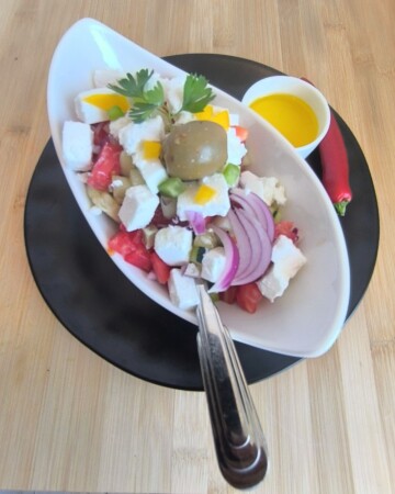 Šopska salata u duguljastoj bijeloj zdjeli s feta sirom, povrćem i maslinom, poslužena s maslinovim uljem i crvenom papričicom na drvenoj podlozi.