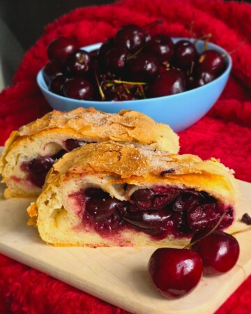 Domaća štrudla od trešanja poslužena na drvenoj dasci, uz svježe trešnje i zdjelu punu voća na crvenoj podlozi.