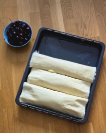 Tri zamotane štrudle od trešanja složene u lim za pečenje, spremne za pećnicu.