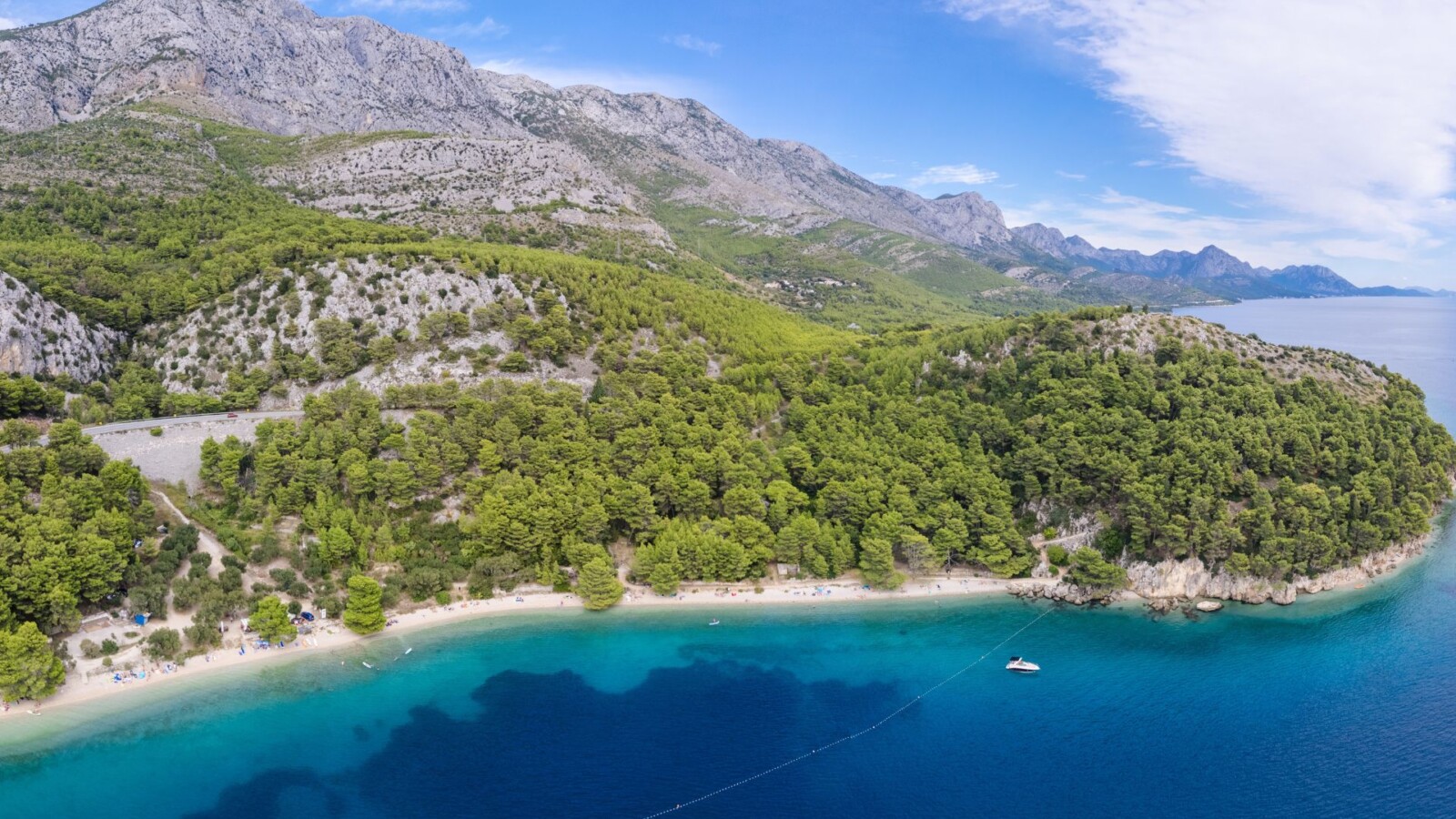 Panoramski pogled na plažu Velika Duba u Živogošću s kristalno čistim morem, gustom borovom šumom i planinskim pejzažem u pozadini.
