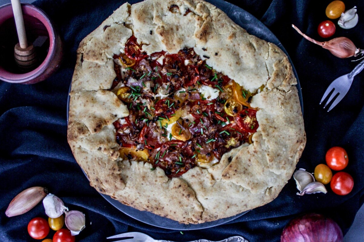 Galette s rajčicama i kozjim sirom na tamnoj pozadini, oko tanjura su svježe rajčice i ljutika