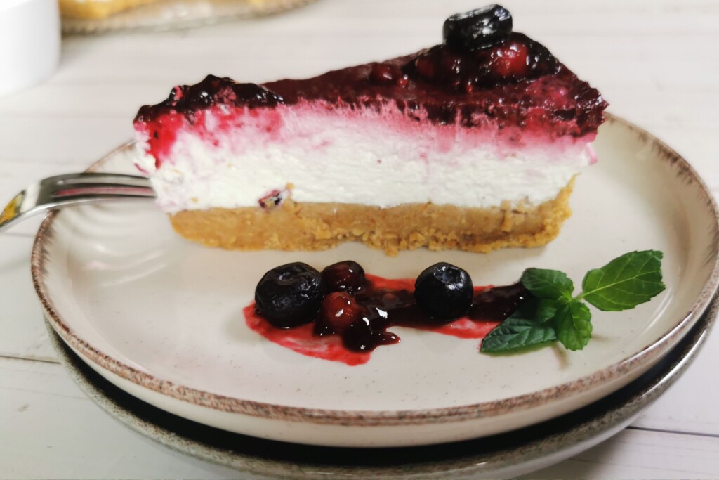 Komad cheesecakea bez pečenja s preljevom od šumskog voća, poslužen na tanjuru uz borovnice i listić svježe mente.