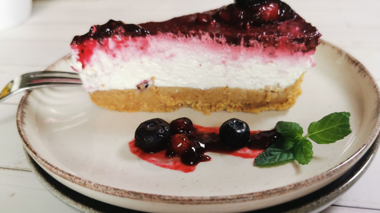 Komad cheesecakea bez pečenja s preljevom od šumskog voća, poslužen na tanjuru uz borovnice i listić svježe mente.