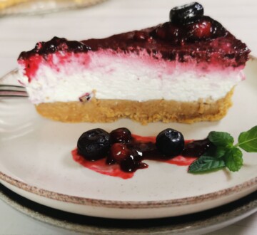 Komad cheesecakea bez pečenja s preljevom od šumskog voća, poslužen na tanjuru uz borovnice i listić svježe mente.