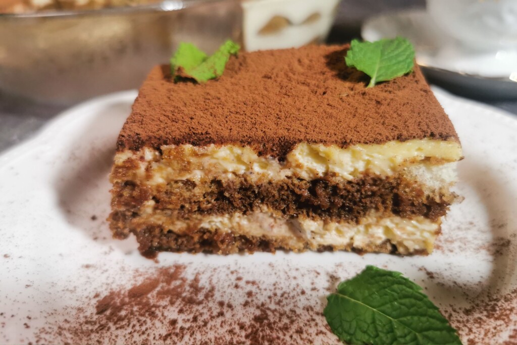 Presjek tiramisua s vidljivim slojevima kreme i piškota, posut kakaom i dekoriran mentom.